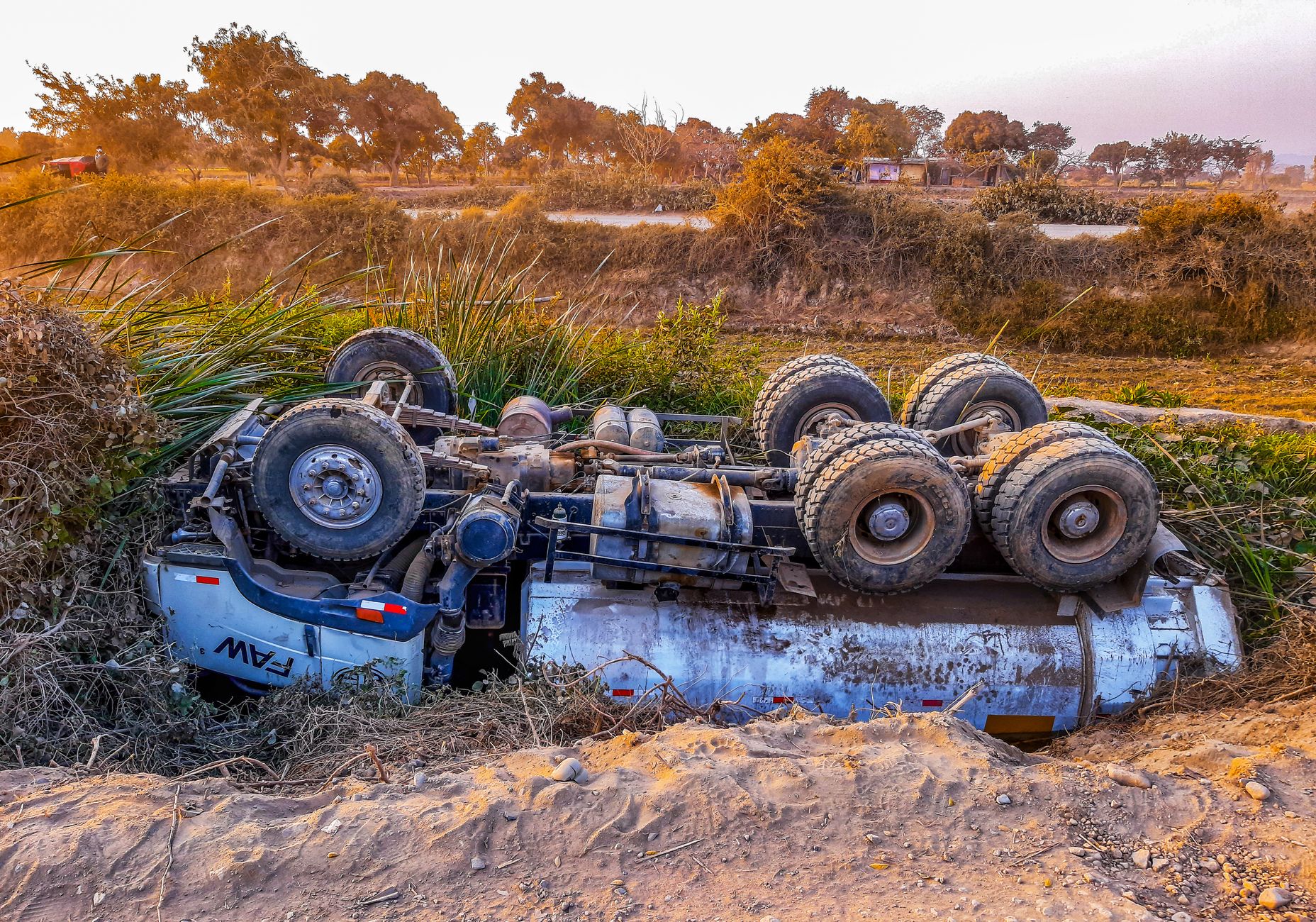 Overturned truck lying on its side beside a rural dirt road during daytime.