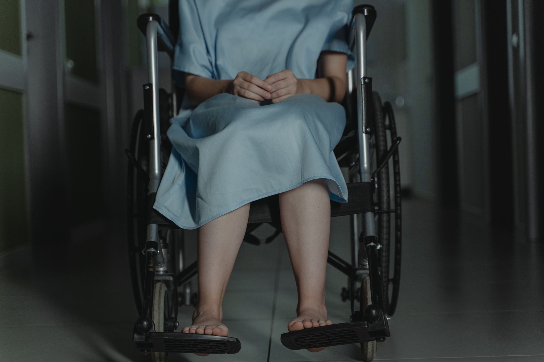 Persona descalza en silla de ruedas con bata de hospital en un pasillo poco iluminado.