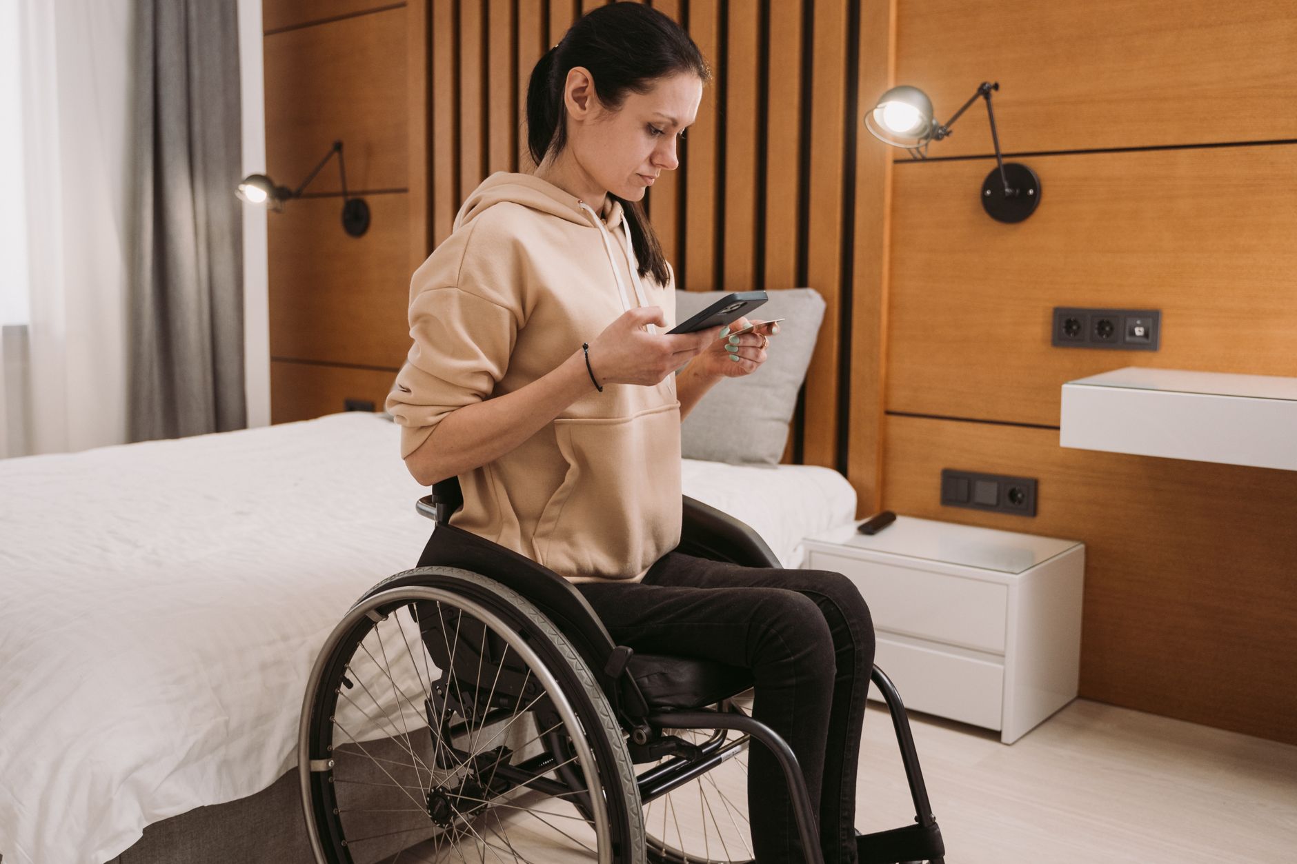 Woman in wheelchair using a smartphone in a wood-paneled bedroom.