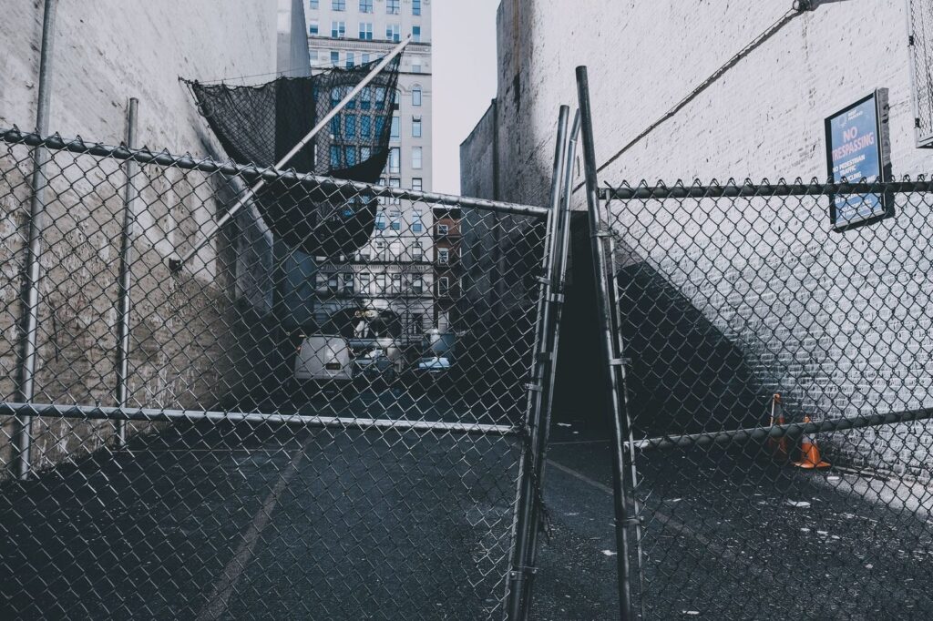 Callejón urbano con cerca de eslabones de cadena y señales de advertencia.