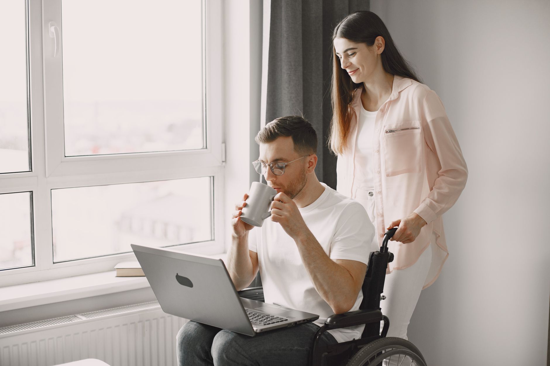 Caregiver assists a man in a wheelchair with coffee and a laptop indoors.