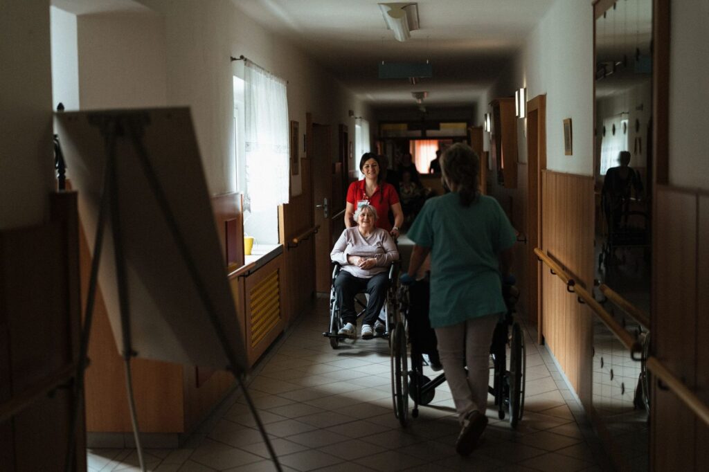 Caregiver pushing elderly woman in wheelchair down a hallway.