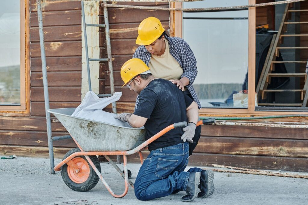 Construction worker kneeling on job site receiving assistance from colleague near wheelbarrow.