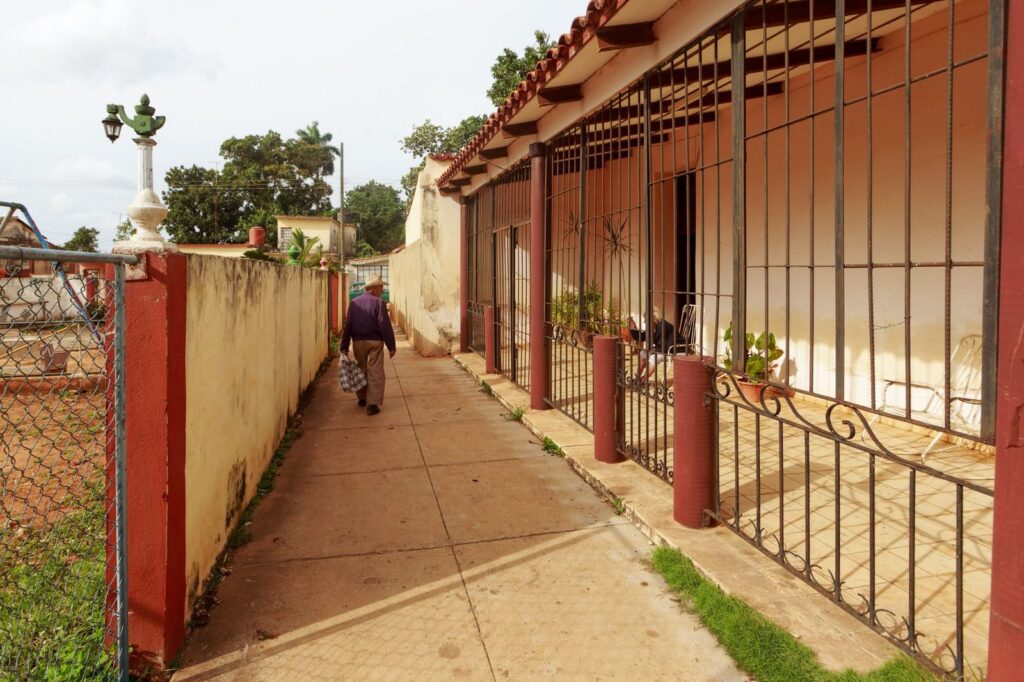 Hombre mayor caminando por sendero junto a cerca de concreto y puerta metálica en zona urbana.