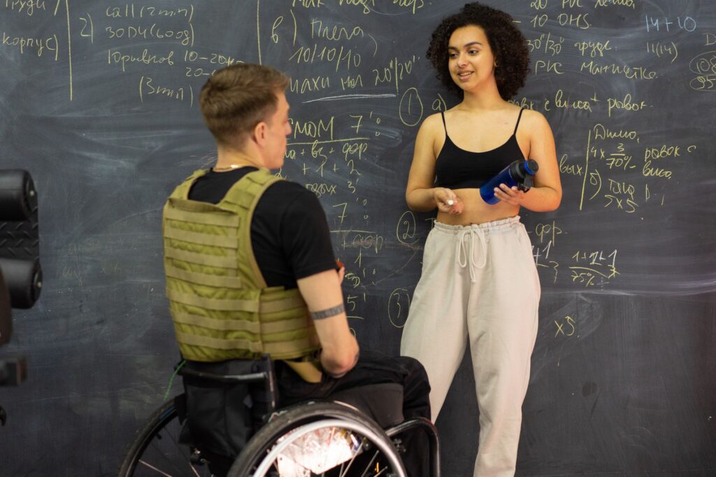 Man in a wheelchair and a woman reviewing a fitness plan on a chalkboard in a gym.