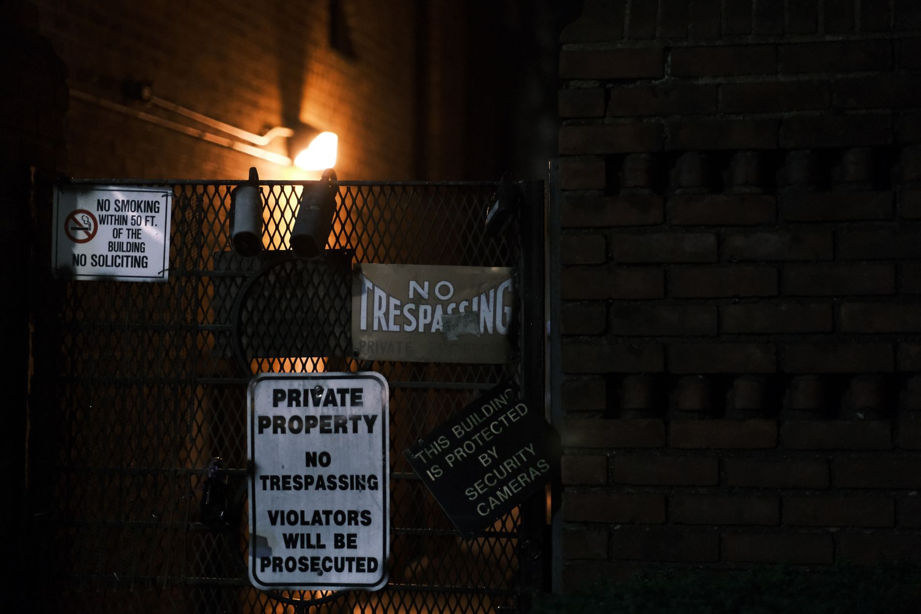 Multiple warning signs posted on a brick wall and gate in a dark urban setting.