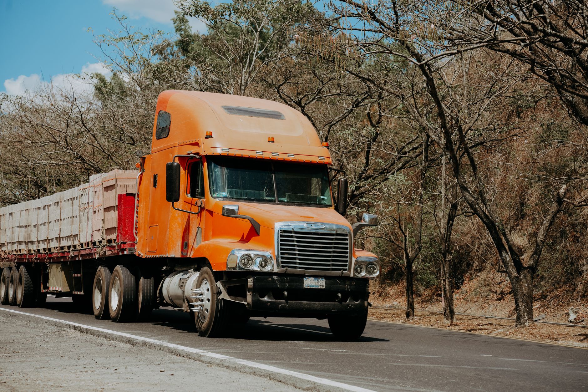 Orange Freightliner semi-truck driving on a highway through a dry, scenic landscape.