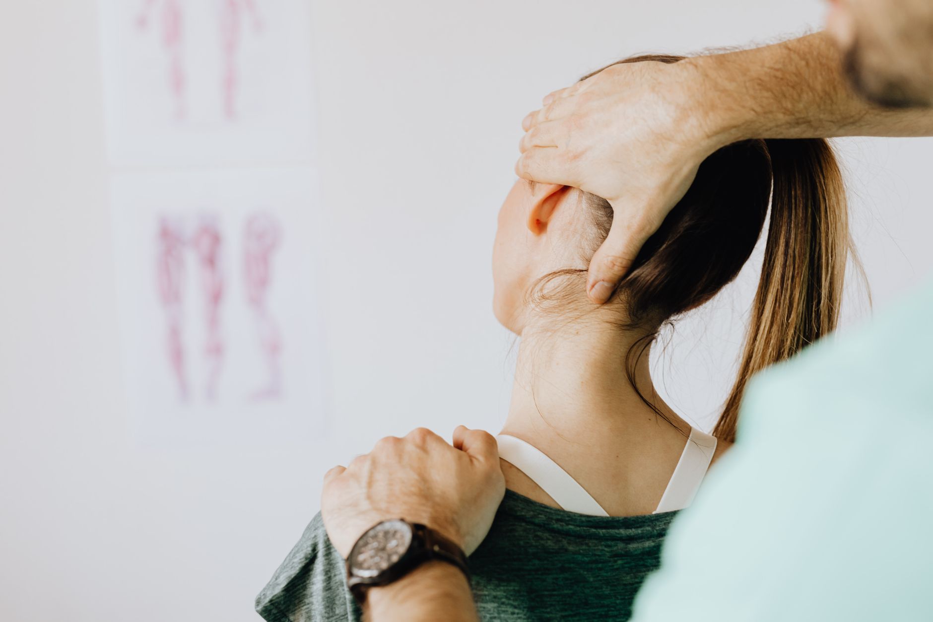 Orthopedist examines woman's neck in office with anatomy drawings on the wall behind them.