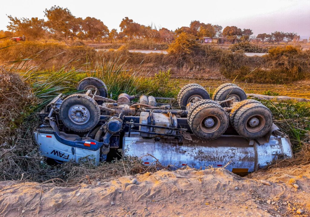Overturned truck lying on its side beside a rural dirt road during daytime.