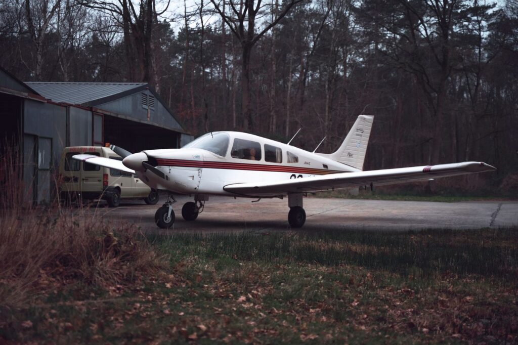 White private aircraft parked on tree-lined airstrip beside metal hangar in forest.