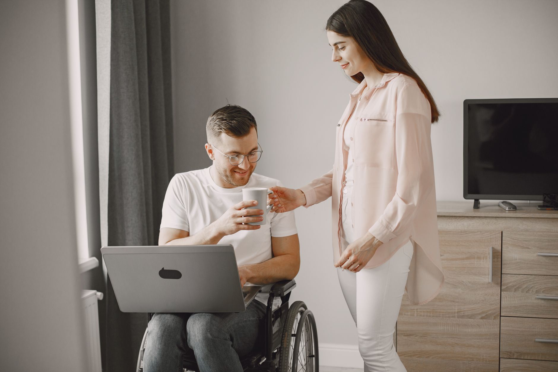 Woman hands a cup to a smiling man using a wheelchair with a laptop on his lap.