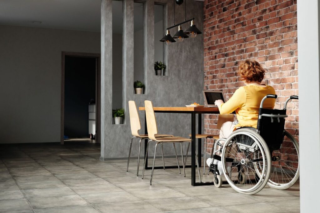 Woman in wheelchair using laptop at home with brick wall background.