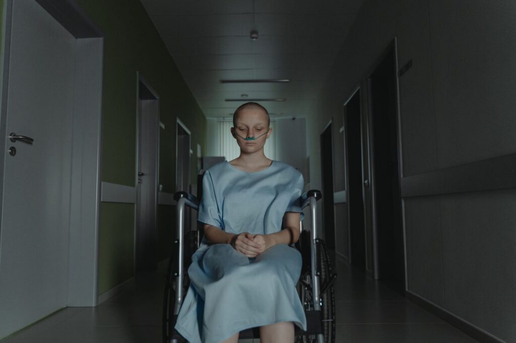 Woman in wheelchair wearing hospital gown sitting alone in dimly lit hospital corridor.