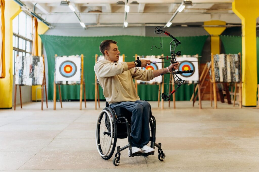 Young man in wheelchair aiming bow and arrow at target indoors.