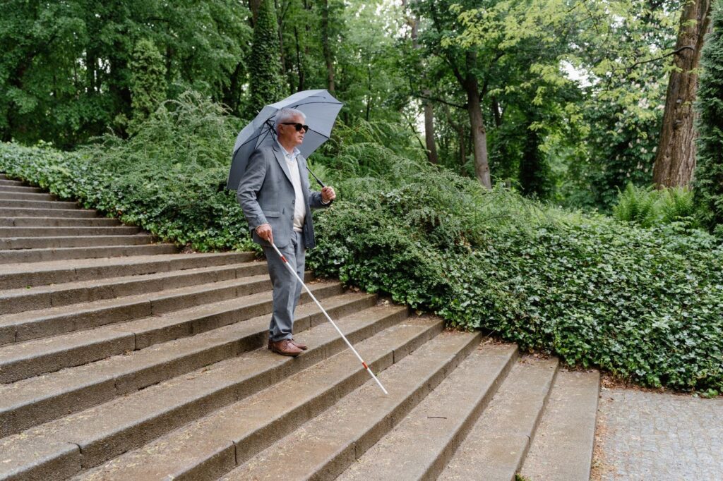 Blind man with white cane navigating outdoor steps during rain.