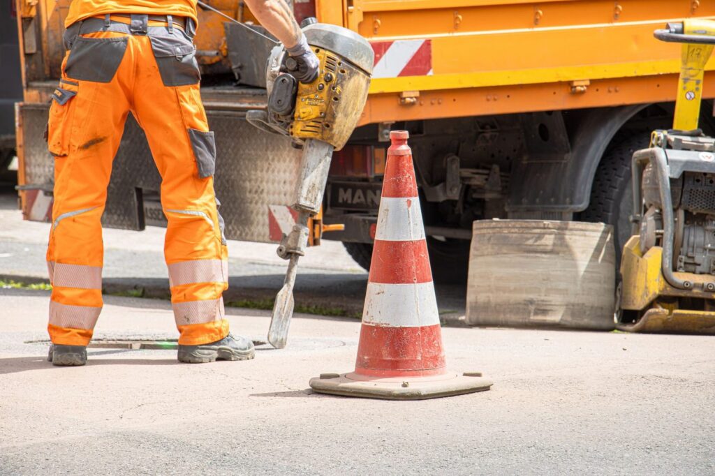 Construction worker operating jackhammer near traffic cone on city street.