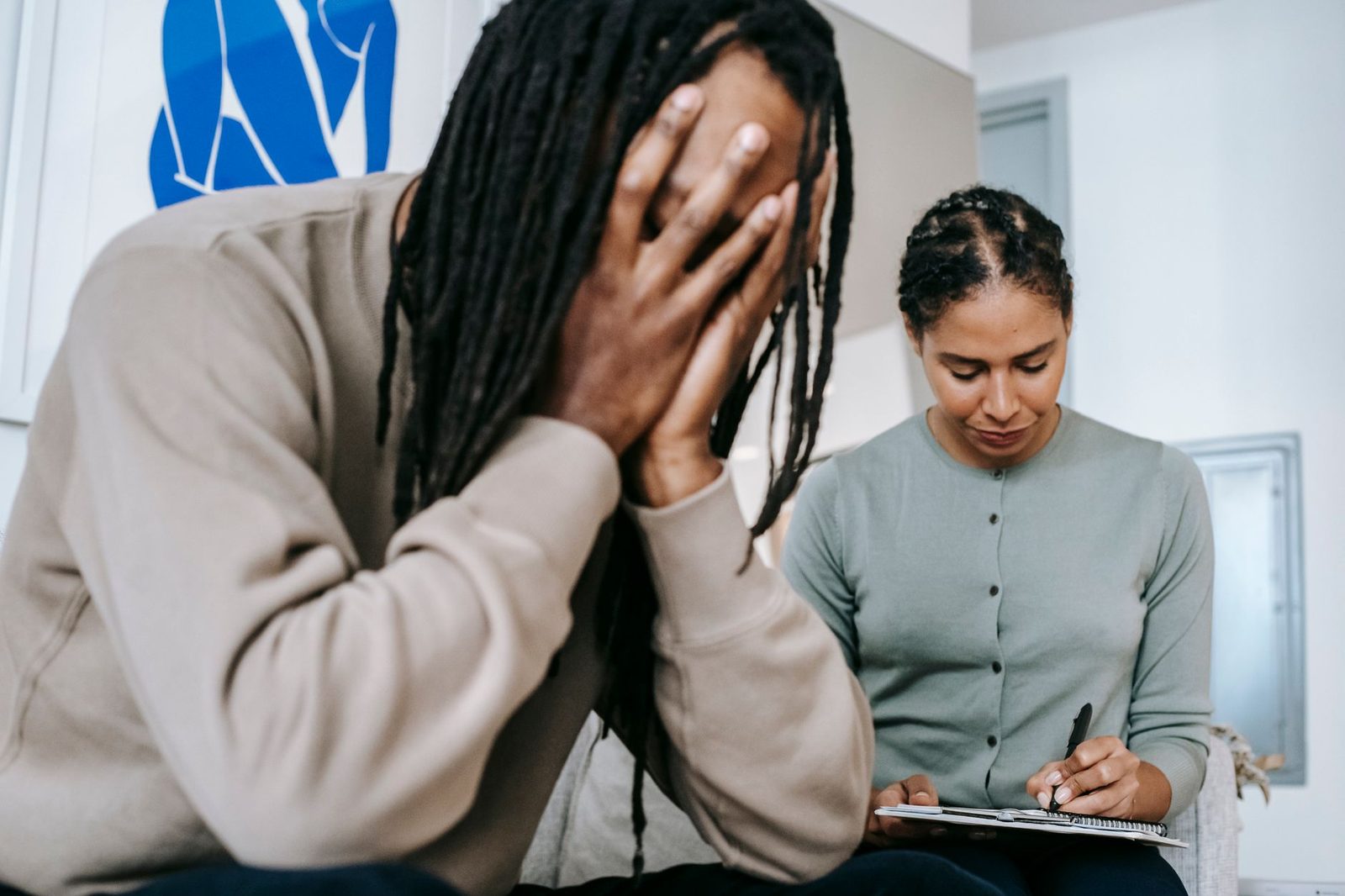 Hombre afroamericano con rastas cubriéndose la cara mientras mujer hispana toma notas en sesión de terapia.