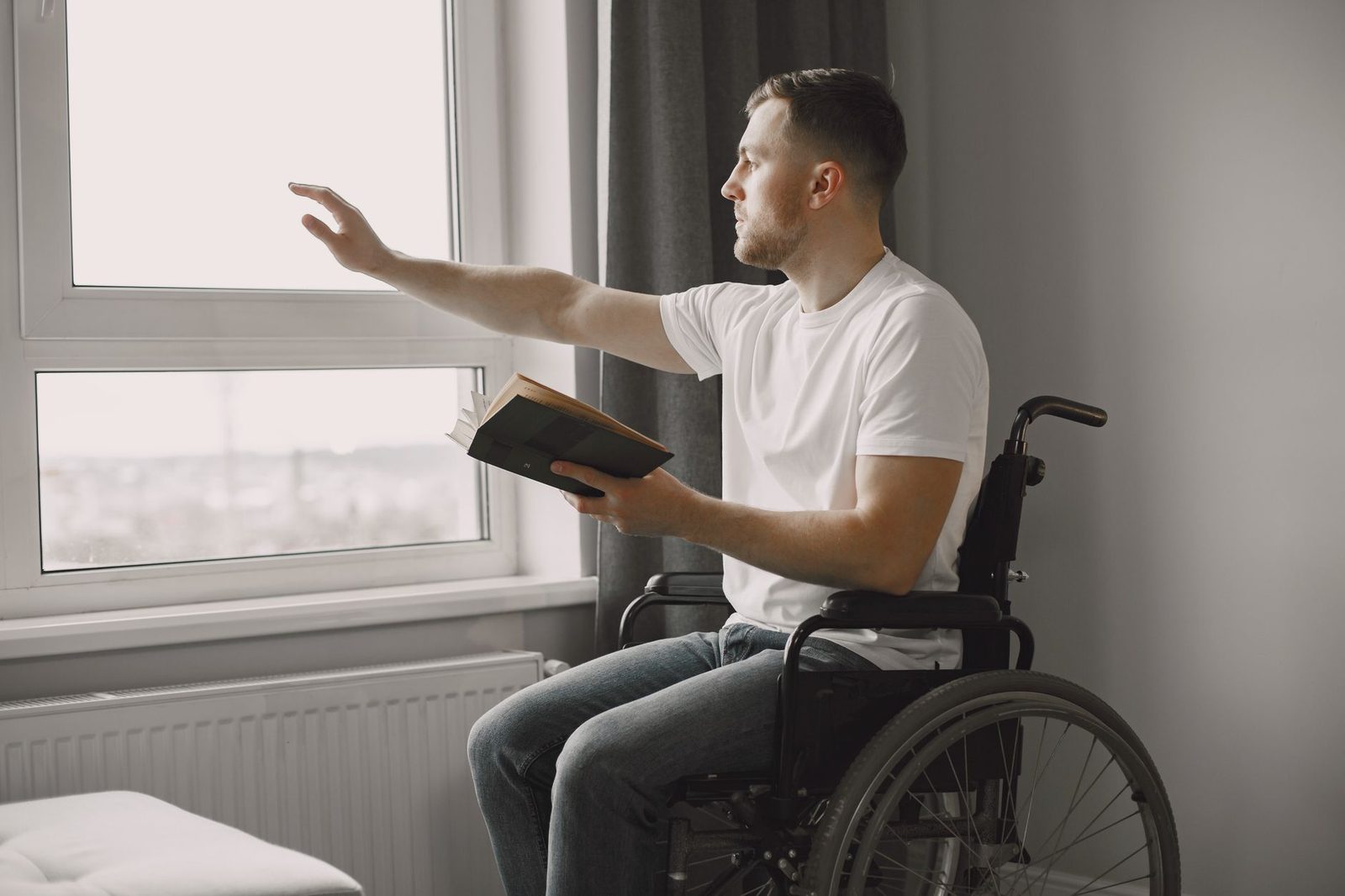 Man in wheelchair reading a book by a window indoors.