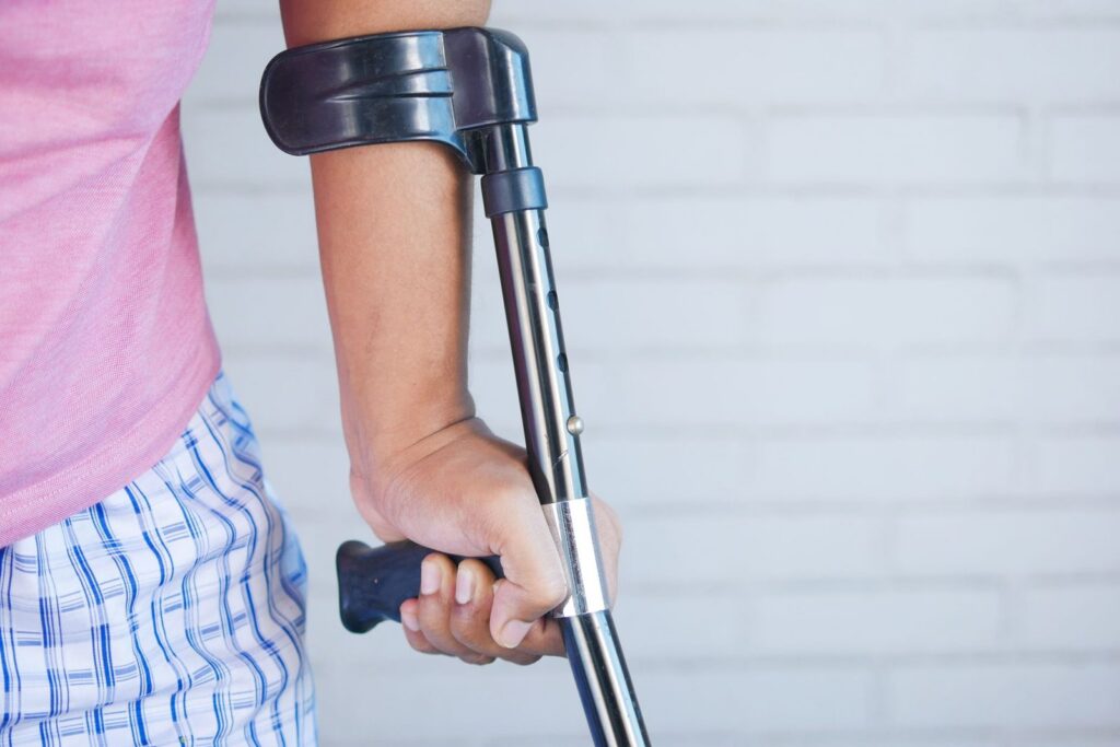 Person in pink shirt using crutch for support against brick wall.