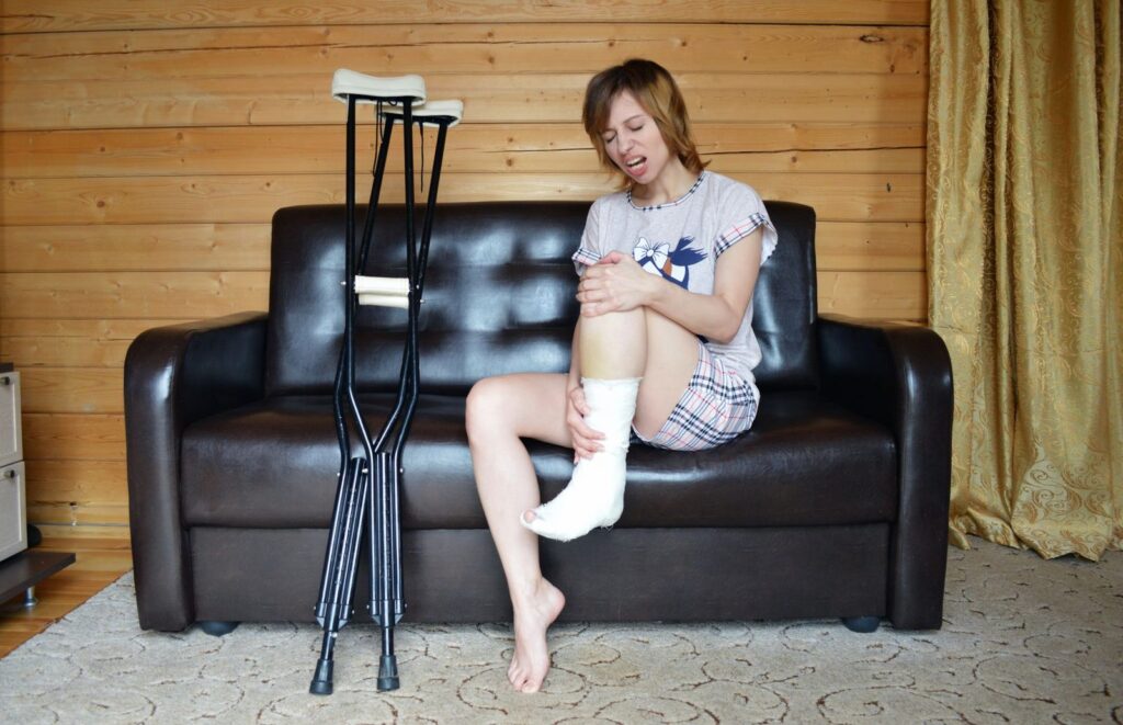 Woman holding her leg in pain while sitting on couch with crutches nearby.