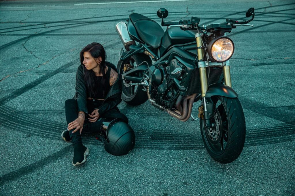 Woman in black clothing sits beside motorcycle on road with skid marks.