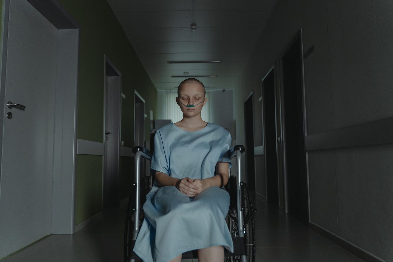 Woman in hospital gown sitting in wheelchair in dimly lit corridor.
