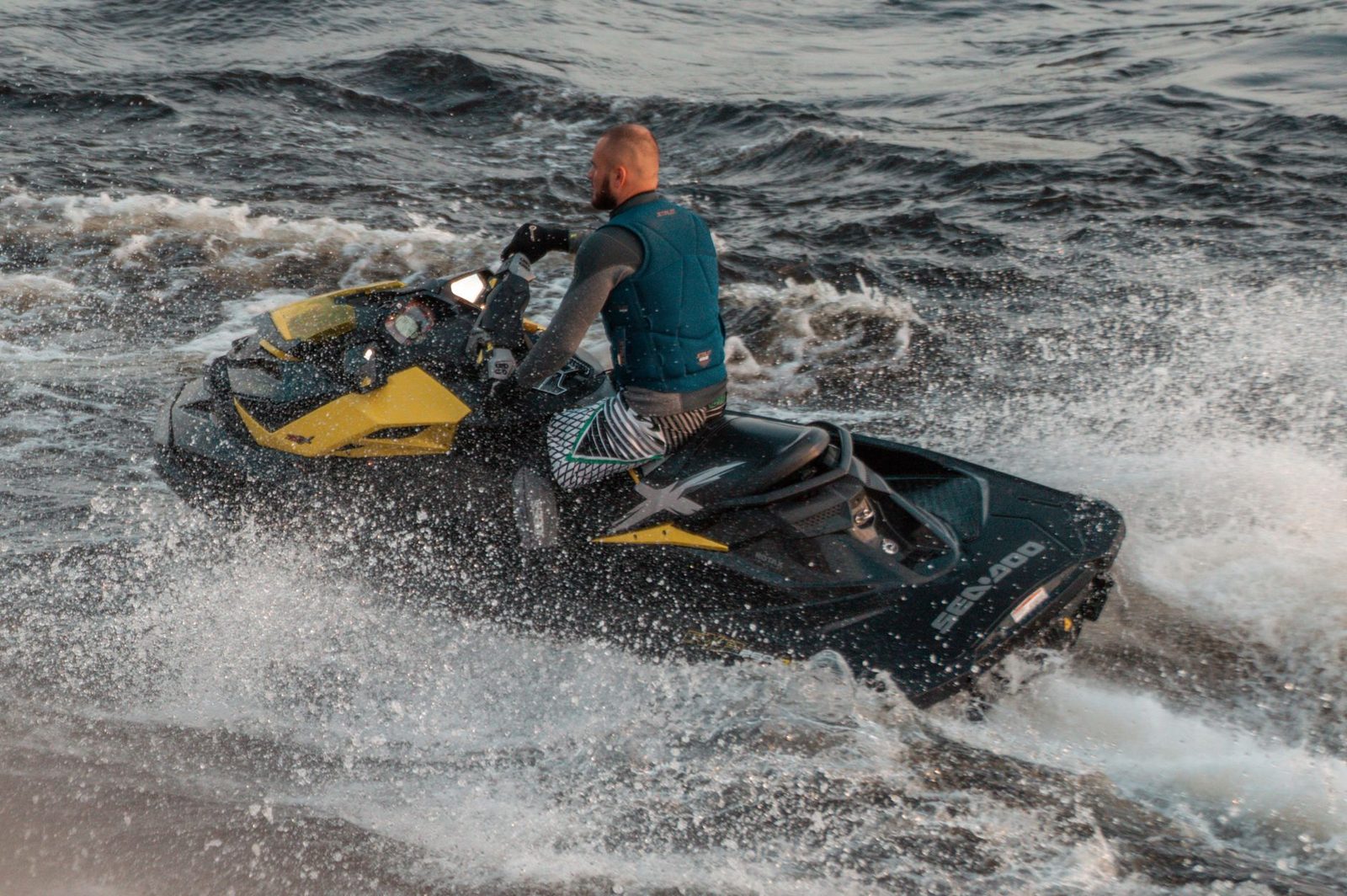 A man in a life vest rides a jet ski across choppy water, throwing up dramatic spray.