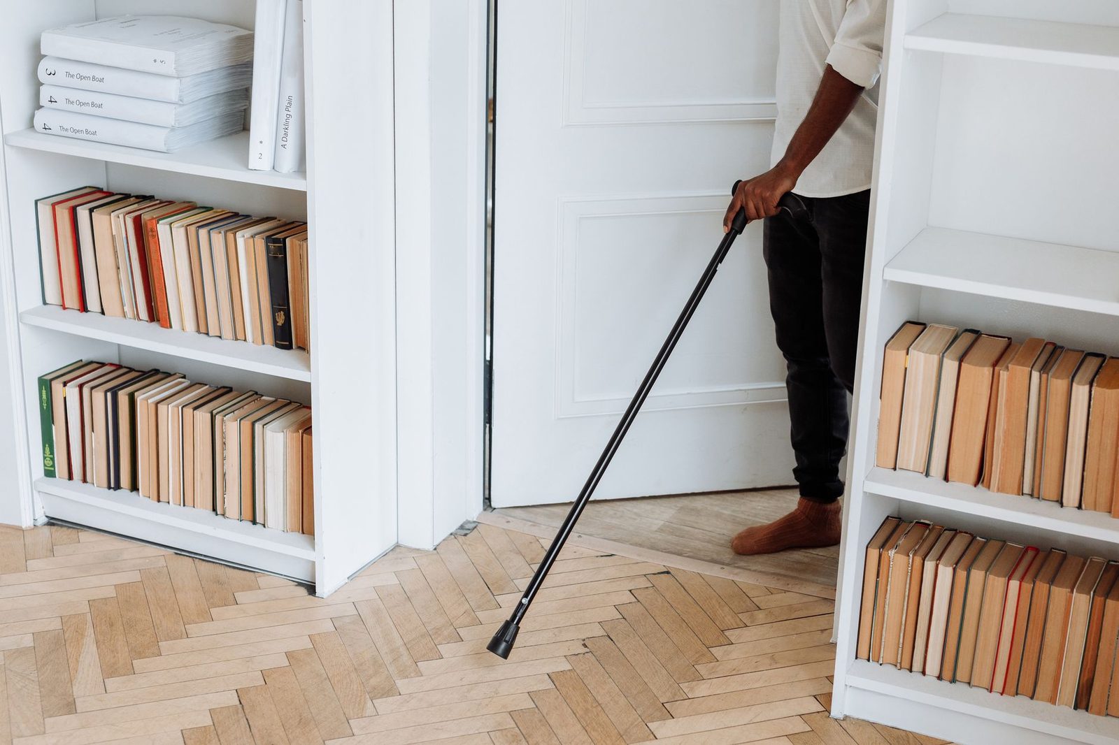 A person with a walking cane moves across a room lined with bookshelves.