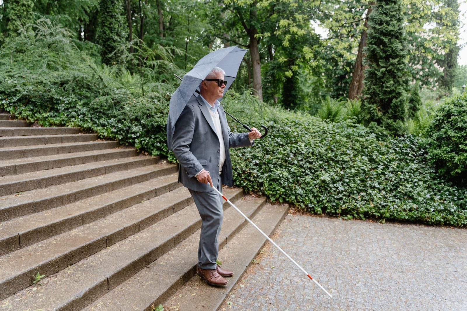 A senior man with a visual impairment walks through a green park, using a white cane and holding an umbrella.