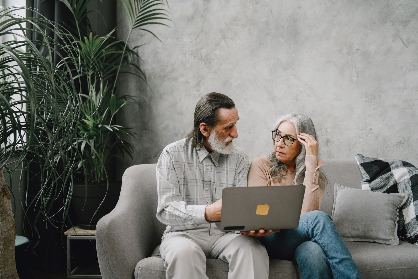 Elderly couple talking on a sofa at home while looking at a laptop, with houseplants nearby.