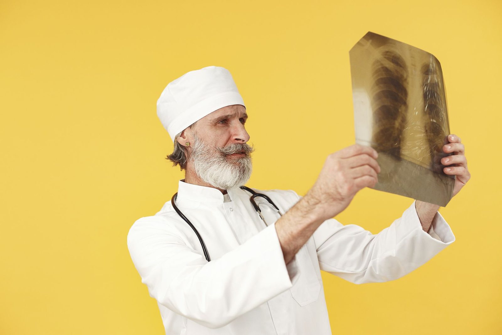 Elderly doctor in a white coat examining a chest X-ray against a bright yellow background.