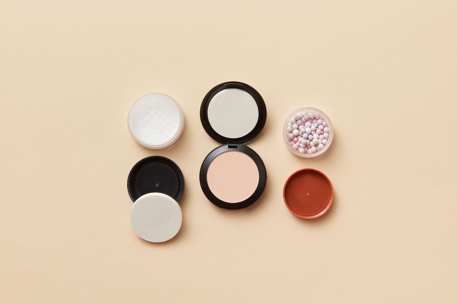 Vista cenital de envases de maquillaje variados y borlas de polvo sobre una superficie beige.