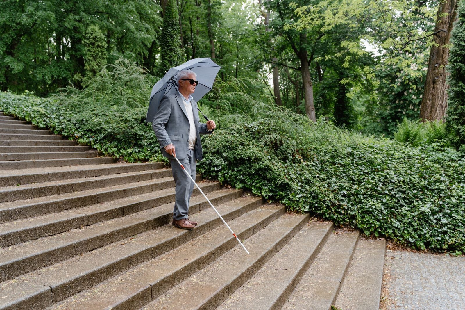 Hombre con bastón blanco desciende escaleras exteriores bajo lluvia.