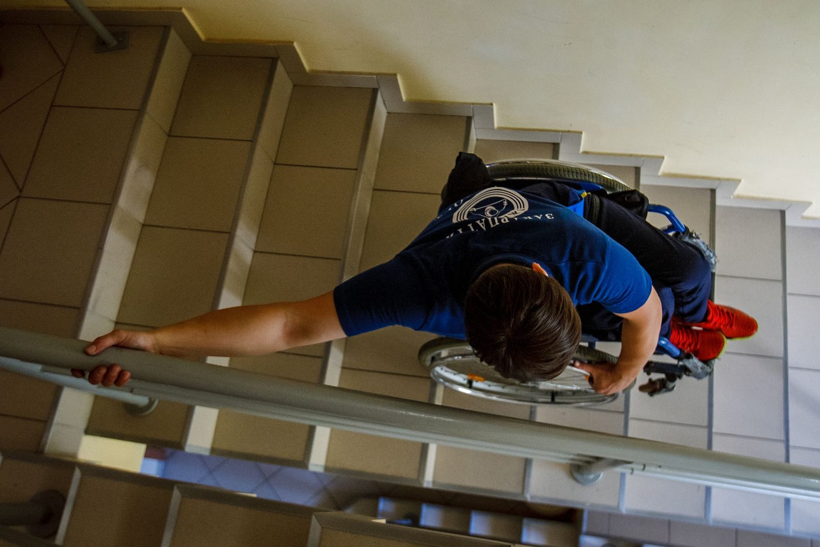 Person in wheelchair descending indoor stairs while holding railings.