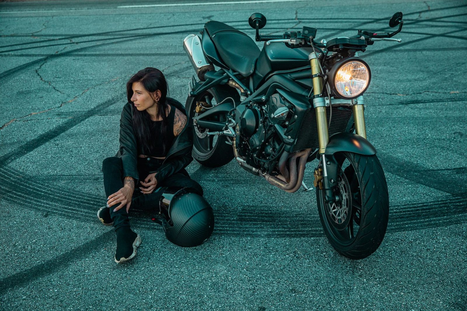 Woman in black clothing sits beside motorcycle on road with skid marks.