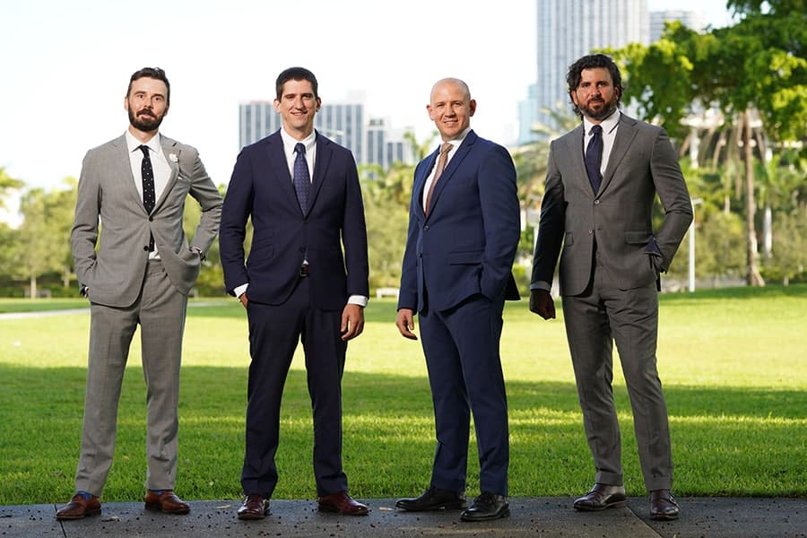 Four smiling men in suits outdoors.